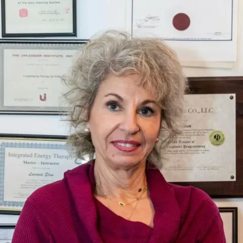 Luciana Stan in front of her framed certificates, including Upledger Institute CranioSacral Therapy, Integrated Energy Therapy Master-Instructor and NLP Trainer