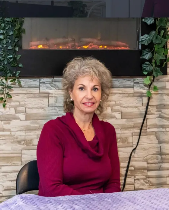 Luciana Stan seated in her treatment room, beside the treatment table, with a fireplace and plants behind her
